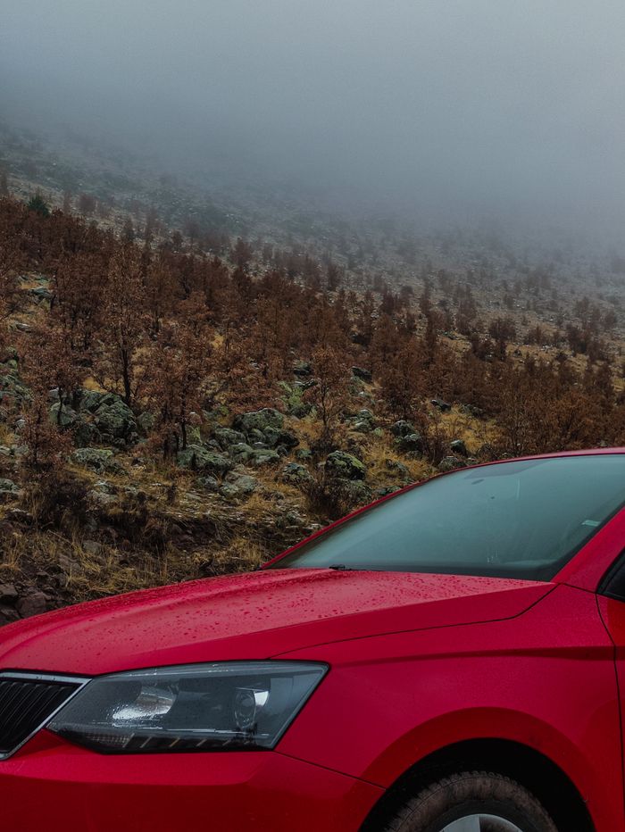 A vibrant red car parked against a misty mountainous backdrop with autumn foliage.